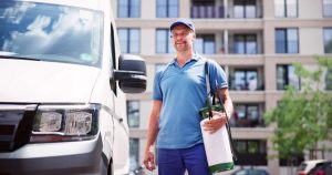 A man in a blue uniform and cap stands beside a white van, holding a large spray canister for home insect removal. The background features a modern apartment building with multiple balconies and some greenery.