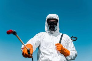 A person wearing a protective suit and mask aims a spray nozzle forward against a clear blue sky. With orange gloves on, they appear ready for serious pest control or bed bug removal tasks, ensuring safety while managing hazardous materials effectively.