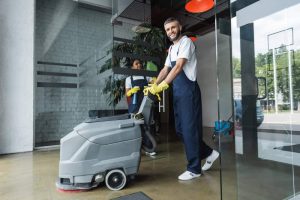 A person smiling while skillfully operating a floor cleaning machine in a modern indoor space with large glass doors, reminiscent of a pest control expert tackling home insect removal with precision. Another individual is visible in the background, both donning uniforms and gloves.