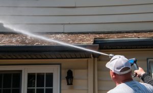A person is pressure washing the shingles of a house roof. They are wearing a white cap and holding a power washer, directing a strong stream of water upwards. The house has beige siding and a lamp next to a window.
