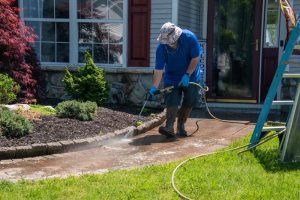 A person wearing a hat, gloves, and boots is pressure washing a brick pathway in front of a house. A ladder is visible on the grass, and a garden with small shrubs lines the path. The front door has a "Welcome" sign.