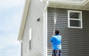 A man wearing a blue shirt is pressure washing the side of a house with gray siding. The hose shoots water towards the upper part of the building, spraying it clean. The sky is partially visible and appears clear.