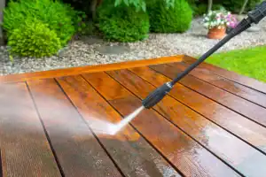 A pressure washer sprays water onto a wooden deck, cleaning the surface. Green bushes, gravel, and potted flowers are visible in the background, suggesting an outdoor garden setting—perfect for those seeking pest control in Franklin, NJ.