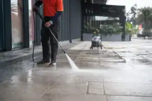 A person wearing boots and an orange shirt uses a pressure washer to clean a large outdoor pavement area near a building, ensuring it stays spotless for pest control in Franklin, NJ.