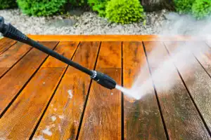A pressure washer sprays water onto a wooden deck, cleaning the surface. The left side looks cleaner and brighter than the right, showing the difference after washing. Green bushes are visible in the background, perfect for pest control in Franklin, NJ.